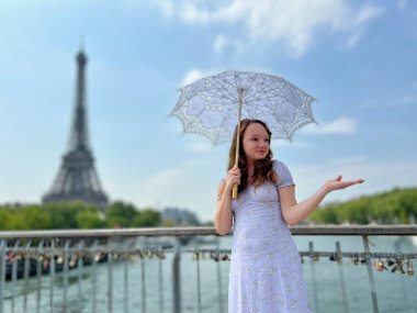 Yazlık elbiseli güzel bir kız dantel bir şemsiyeyle Eyfel Kulesi 'nin arka planında duruyor, elini kaldırdı ve sanki seyahat acentesinde bir yer varmış gibi havalanıyor.