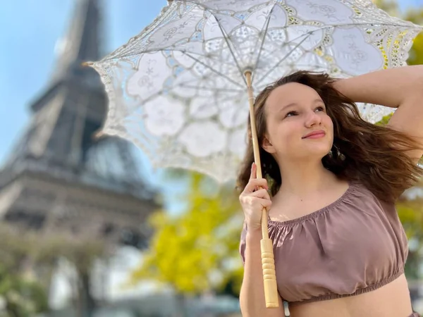 一个穿着夏装的年轻漂亮姑娘站在埃菲尔铁塔的后面，手里拿着花边雨伞，她举起手，好像是在那里为旅行社做广告似的 — 图库照片