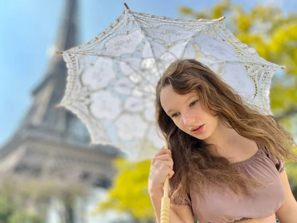 A beautiful young girl in a summer dress stands against the backdrop of the Eiffel Tower with a lace umbrella, she raised her hand and as if taking off there is a place for advertising travel agency — Stock fotografie