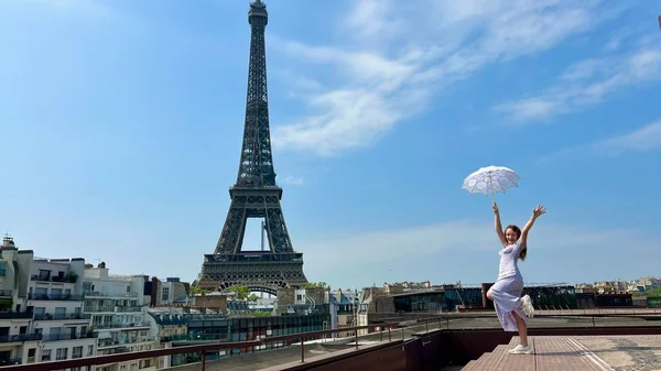 A beautiful young girl in a summer dress stands against the backdrop of the Eiffel Tower with a lace umbrella, she raised her hand and as if taking off there is a place for advertising travel agency — Foto Stock