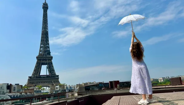 A beautiful young girl in a summer dress stands against the backdrop of the Eiffel Tower with a lace umbrella, she raised her hand and as if taking off there is a place for advertising travel agency — Stock fotografie