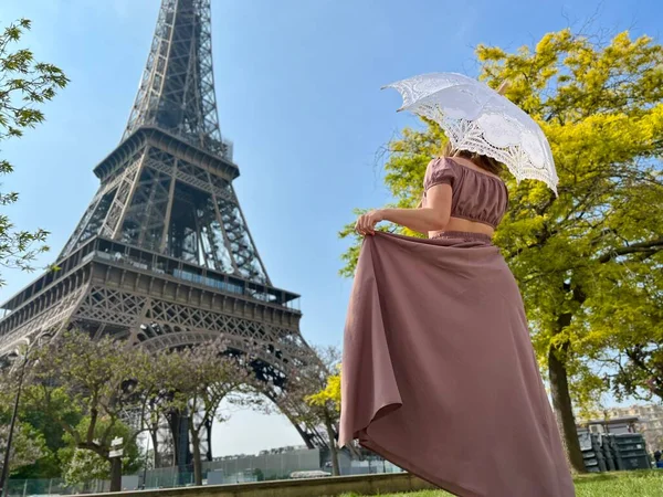 Сзади мы видим девушку в красивом длинном коричневом платье в стиле ретро с зонтиком, который идет к Эйфелевой башне держа платье одной рукой — стоковое фото