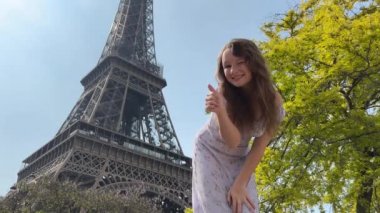 a beautiful teenage girl looks into the frame leaning against the backdrop of the Eiffel Tower She smiles and seems to be showing come here great advertisement for a trip to Paris