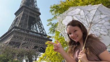 a beautiful teenage girl looks into the frame leaning against the backdrop of the Eiffel Tower She smiles and seems to be showing come here great advertisement for a trip to Paris