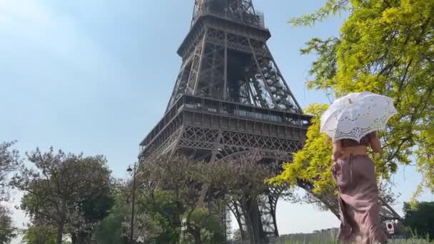 A girl in a long dress with an openwork umbrella goes to the eiffel tower with her back to the camera can be used for advertising text for the interior very beautiful slow motion video — Stockvideo