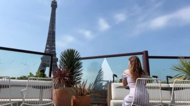 Genç bir kız Eiffel Kulesi 'ne tepeden bakan bir restoranda sırtı dönük oturuyor, rüzgar saçlarını savuruyor. Restoranların ve tur operatörlerinin reklamları ve metinleri var..