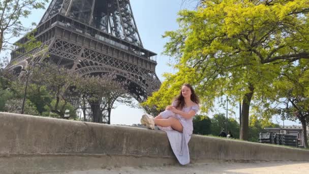 A beautiful teenage girl sits near the Eiffel Tower and a green tree and waves her hands with joy, she seems to be dancing and having fun there is a place for text about travel joy partying — Stockvideo