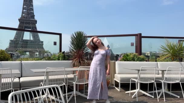 A beautiful teenager girl in a long blue dress stands against the backdrop of the Eiffel Tower in a restaurant, she straightens her hair and leaves the dress is blown by the wind of space for text — Vídeo de stock