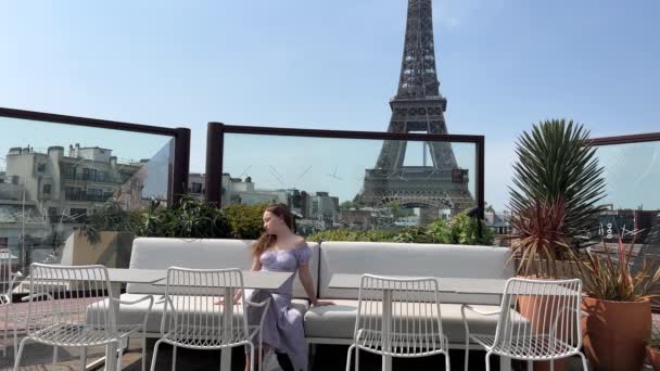 Egy szép hajú lány ül egy hosszú, gyönyörű ruhában az Eiffel-torony háta mögött, szöveges szöveggel egy étteremben, kilátással az Eiffel-toronyra. Hátradől az ülésre. — Stock videók