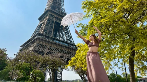 Ein schönes junges Mädchen tanzt und dreht in der Nähe des Eiffelturms mit einem durchbrochenen Sonnenschirm, über den sie sich freut, der für jede Werbung für Freude und Glück verwendet werden kann — Stockfoto