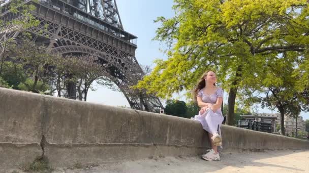 Ung vacker flicka i en blå klänning sitter nära Eiffeltornet nära ett grönt träd hon rätar ut sina flätor ser sig omkring hon är glad och mycket glad — Stockvideo