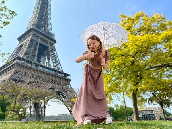 A cheerful teen girl stands against the backdrop of the Eiffel Tower in a beautiful dress, she raised her hand, you can advertise a travel agency trip to Paris France — Zdjęcie stockowe