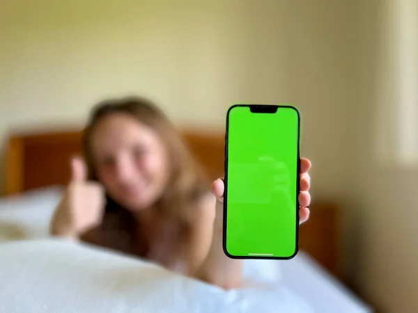 Het meisje lacht ligt in haar bed en toont de telefoon met een groen scherm kan worden gebruikt om seizoensgebonden allergieën of verkoudheden adverteren Wanneer het meisje al is hersteld — Stockfoto