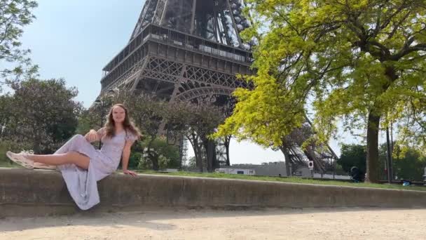 A beautiful teenage girl sits near the Eiffel Tower and a green tree and waves her hands with joy, she seems to be dancing and having fun there is a place for text about travel joy partying — стокове відео