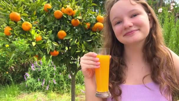 Девочка-подросток стоит со стаканом апельсинового сока на фоне цитрусовых деревьев мандаринов или апельсинов, висящих на дереве. Она улыбается и смотрит в кадр, который можно использовать для рекламы соков — стоковое видео