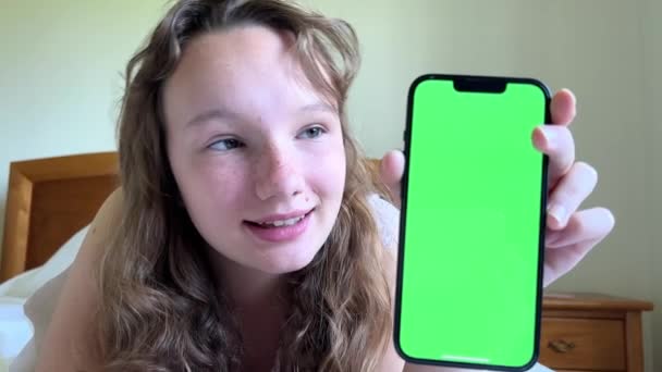 La chica sonríe se encuentra en su cama y muestra el teléfono con una pantalla verde se puede utilizar para anunciar alergias estacionales o resfriados Cuando la chica ya se ha recuperado — Vídeos de Stock