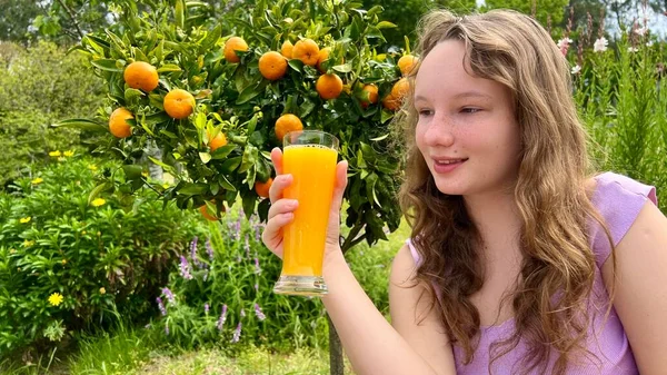A menina bebe suco de laranja no fundo de uma árvore de tangerina, pode ser manga de tangerina de suco de laranja ela bebe avidamente e realmente gosta do suco delicioso em todos os lugares verdes e verão — Fotografia de Stock