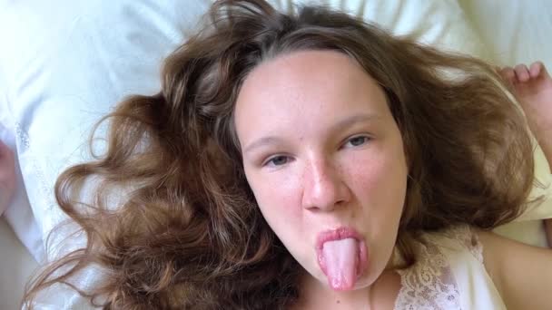 Adolescente menina mostra sua língua no quadro ela se encontra em um travesseiro branco — Vídeo de Stock