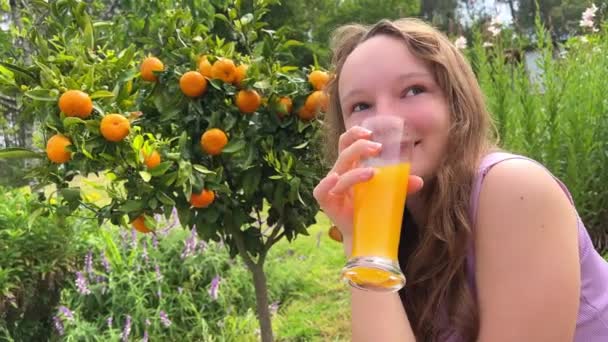 En tonåring flicka på bakgrunden av ett apelsinträd i hennes händer med ett glas apelsinjuice blinkar in i ramen och gör ansikten — Stockvideo