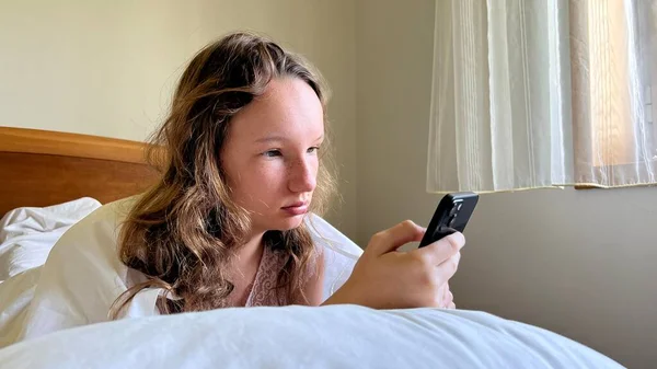 Девушка в белой рубашке подросток лежит на белой кровати в руках она держит черный iPhone 13 Она смотрит на экран спокойно поднимая руки — стоковое фото