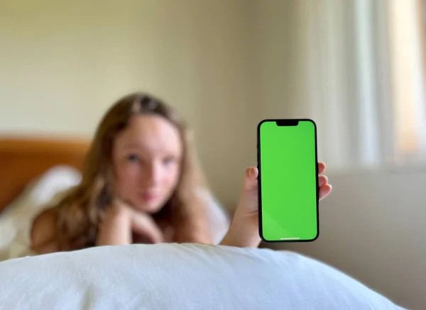 Dívka v bílé košili teenager leží na bílé posteli v rukou drží černý iPhone 13 Dívá se na obrazovku klidně zvedá ruce — Stock fotografie
