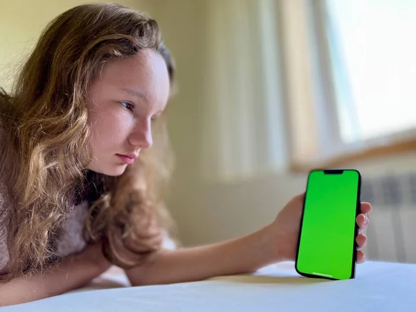 Uma menina em uma camisa branca um adolescente encontra-se em uma cama branca em suas mãos ela segura um iPhone preto 13 Ela olha para a tela calmamente levantando as mãos — Fotografia de Stock