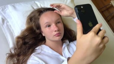 Beyaz gömlekli bir kız elinde beyaz bir yatakta uzanmış siyah bir iPhone 13 'ü tutuyor sakince ekrana bakıyor ve ellerini kaldırıyor.