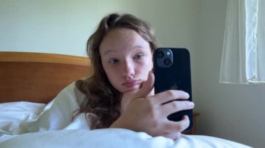 Beyaz gömlekli bir kız elinde beyaz bir yatakta uzanmış siyah bir iPhone 13 'ü tutuyor sakince ekrana bakıyor ve ellerini kaldırıyor.
