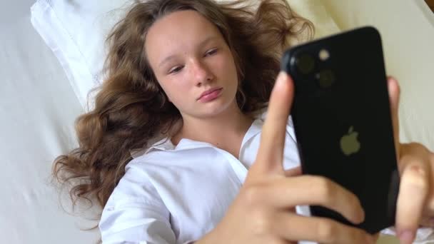 Dívka v bílé košili teenager leží na bílé posteli v rukou drží černý iPhone 13 Dívá se na obrazovku klidně zvedá ruce — Stock video