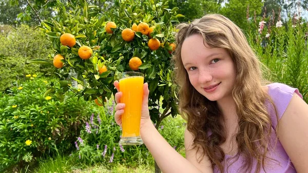 오렌지 주스를 청록색 나무의 배경에 놓고 마시는 여자 애는 오렌지 주스를 톡 쏘는 망고를 먹을 수있고 설탕물을 탐욕스럽게 마실 수있다. — 스톡 사진