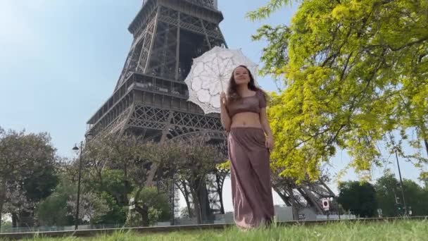 Joven hermosa chica en un vestido azul se sienta cerca de la Torre Eiffel cerca de un árbol verde ella endereza sus trenzas mira a su alrededor ella es feliz y muy feliz — Vídeos de Stock