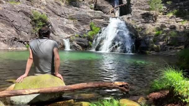 Ciemnowłosy mężczyzna siedzi plecami do kamery i patrzy na wodospad i wodę, którą siedzi na kamieniu ubrany w siwą koszulkę — Wideo stockowe