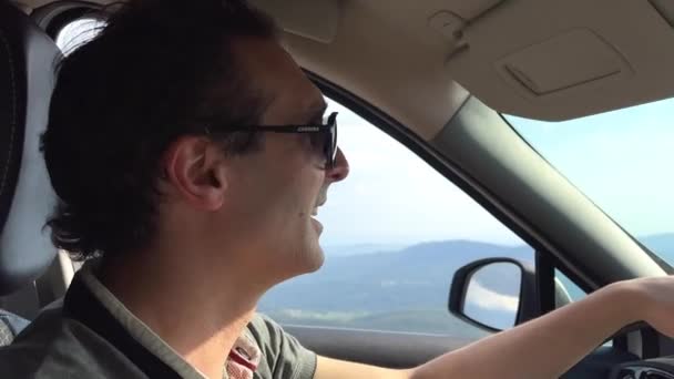 Чоловік у сонцезахисних окулярах водить машину. У нього гарне італійське обличчя. Він вказує на відстань і посміхається за вікном гірської скелі і неба. — стокове відео