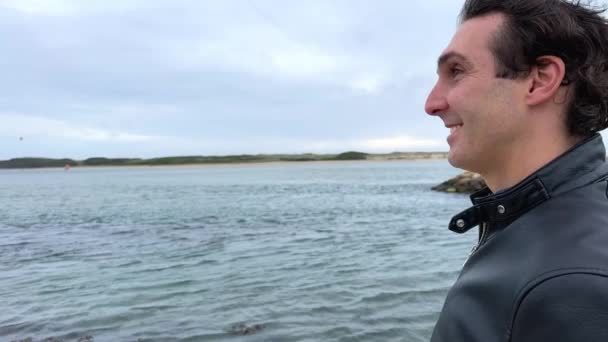 Um homem francês bonito de casaco preto fica nas margens do Oceano Atlântico, tem cabelo preto, uma jaqueta de couro e voa com as mãos para os lados publicidade viagens — Vídeo de Stock