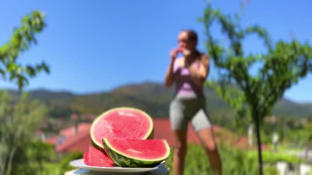 A girl in a summer sports outfit bites a watermelon against the backdrop of mountains she is out of focus in the foreground lies a watermelon — Stock Video