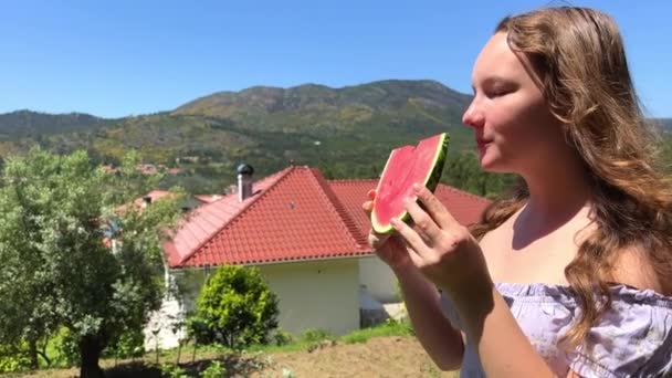 Une jeune fille aux cheveux fluides, une adolescente vêtue d'une robe bleue, debout sur le fond de montagnes et le toit de la maison, elle mord une tranche rouge de pastèque — Video