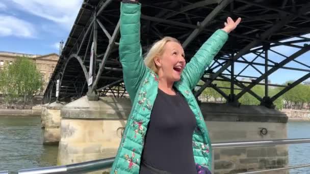 Доросла жінка в зеленій куртці плаває під мостом у Парижі і вигукує, розмахуючи руками, повільно рухаючи відео, вона має велике щастя і радість. — стокове відео