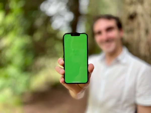 Rozmazané zaostření pouze telefon v ohnisku velmi hezký muž francouzsky italsky nedrží telefon se zeleným kohoutkem stojící v lese v bílé košili a s úsměvem ukazující prst na obrazovce může použít pro — Stock fotografie