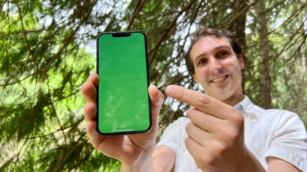英俊潇洒的法国人，意大利佬，没有带着绿色水龙头的手机，穿着白衬衫站在森林里，微笑着在屏幕上显示一个手指可以用来做广告 — 图库照片