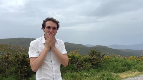 Un hombre guapo de apariencia europea sopla un beso en el marco que mira a la cámara que está en una camisa blanca detrás de él las montañas y el cielo puede ser francés o puede ser español — Vídeos de Stock