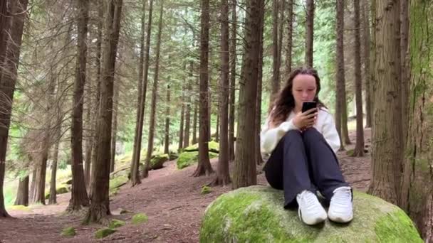 Dívka v bílé košili teenager sedí na velkém kameni v lese ve svých rukou drží černý iPhone 13 Dívá se na obrazovku klidně zvedá ruce — Stock video