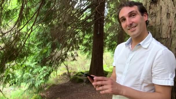 Nagyon jóképű férfi francia olasz nem tartja a telefont egy zöld csap áll az erdőben egy fehér ing és mosolyog mutatja az ujját a képernyőn lehet használni reklám — Stock videók