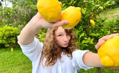 Açık renk saçlı bir kız, tropik bir ülkede limon ağaçlarının arka planında limon hokkabazlığı yapar, beyaz bir gömlek giyer, güler ve eğlenir.