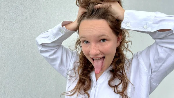白いブラウスに色白の髪をした女の子は陽気な感情を見せ顔を作り喜んで笑い髪をフリルして舌を見せる — ストック写真