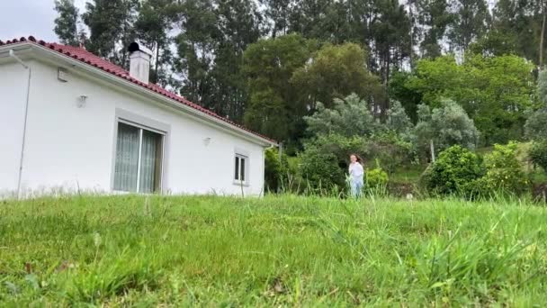 Девушка вся в белом бежит по зеленой траве на фоне белого дома, а затем черный кот пересекает ее путь девушка Стоп не знает, что делать наугад или нет — стоковое видео