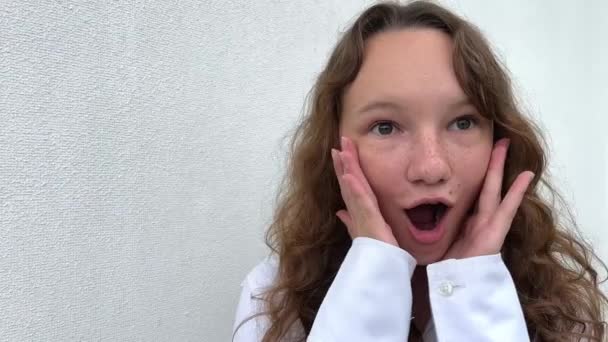 Uma menina em um fundo branco em uma blusa branca, de cabelos claros, mostra diferentes emoções alegres, ela faz rostos, ela ri feliz, ela esfrega o cabelo e mostra sua língua — Vídeo de Stock