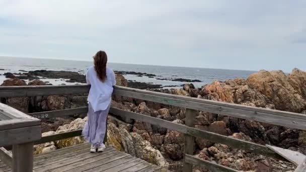 Το κορίτσι περπατάει στο φόντο του Ατλαντικού Ωκεανού, τα μαλλιά της αναπτύσσονται. Είναι ντυμένη με λευκό μπουφάν. Ο καιρός είναι κρύος. βετεράνοι. — Αρχείο Βίντεο