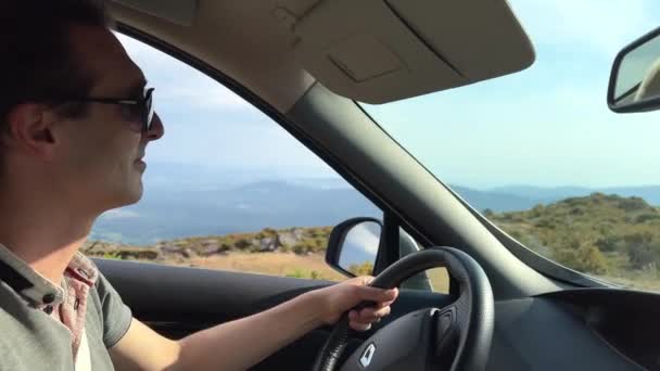 Чоловік у сонцезахисних окулярах водить машину. У нього гарне італійське обличчя. Він вказує на відстань і посміхається за вікном гірської скелі і неба. — стокове відео
