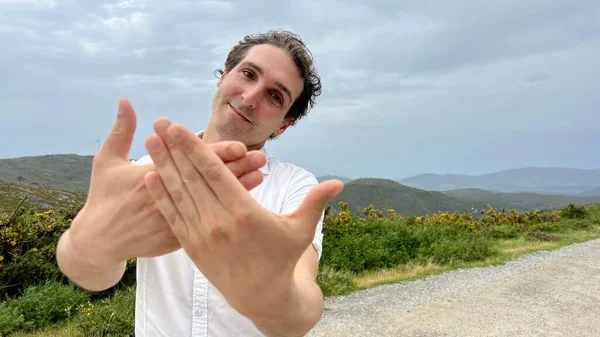 A handsome European man looks at the sky and raises his hands up he is in a white shirt and mountains behind him — Stock Photo, Image