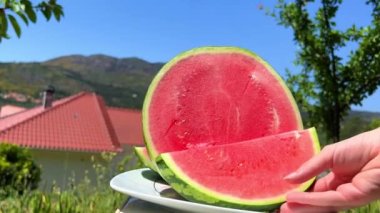 Kesilmiş karpuz, dağların arka planında yatar. Ve tabiatta kadın eli bir parça alır ve tabaktan alır.